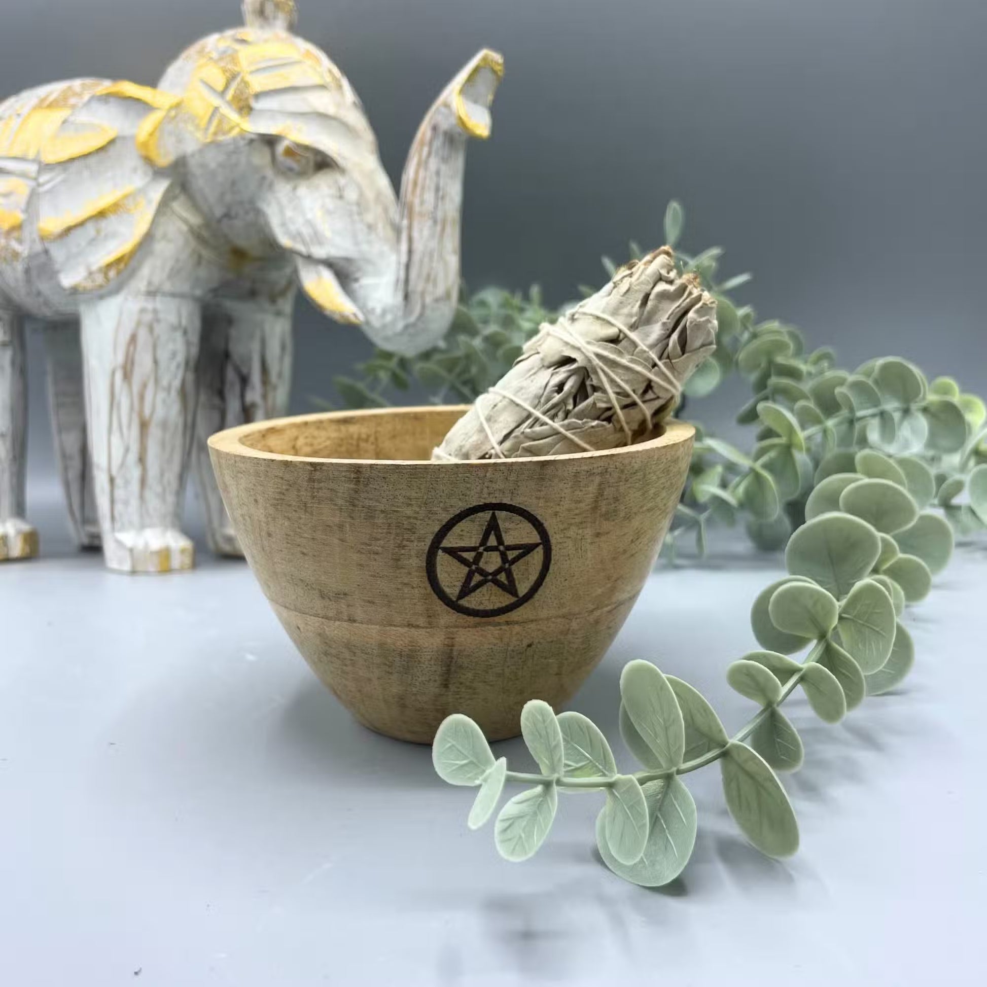 Wooden bowl with a pentagram symbol containing a sage bundle, next to a decorative elephant and eucalyptus leaves on a gray background.