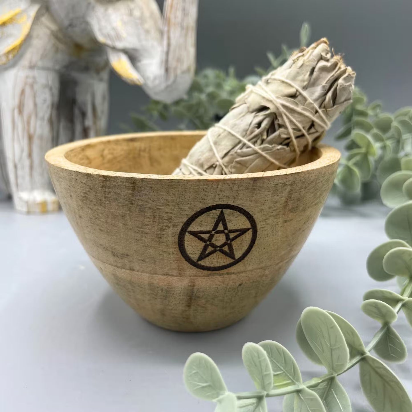 Wooden bowl with a pentagram symbol containing a sage bundle, surrounded by greenery.
