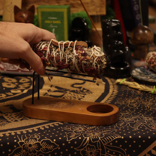 Hand holding a banjara sage smudge stick over a wooden holder on a patterned surface.